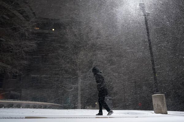 Snow falls past a streetlight. A person in a hooded winter coat leans into the wind.
